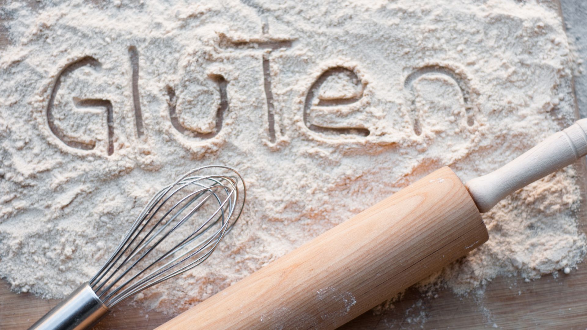 Napis gluten napisany na mące, a obok niego przedmioty do wyrabiania ciasta.